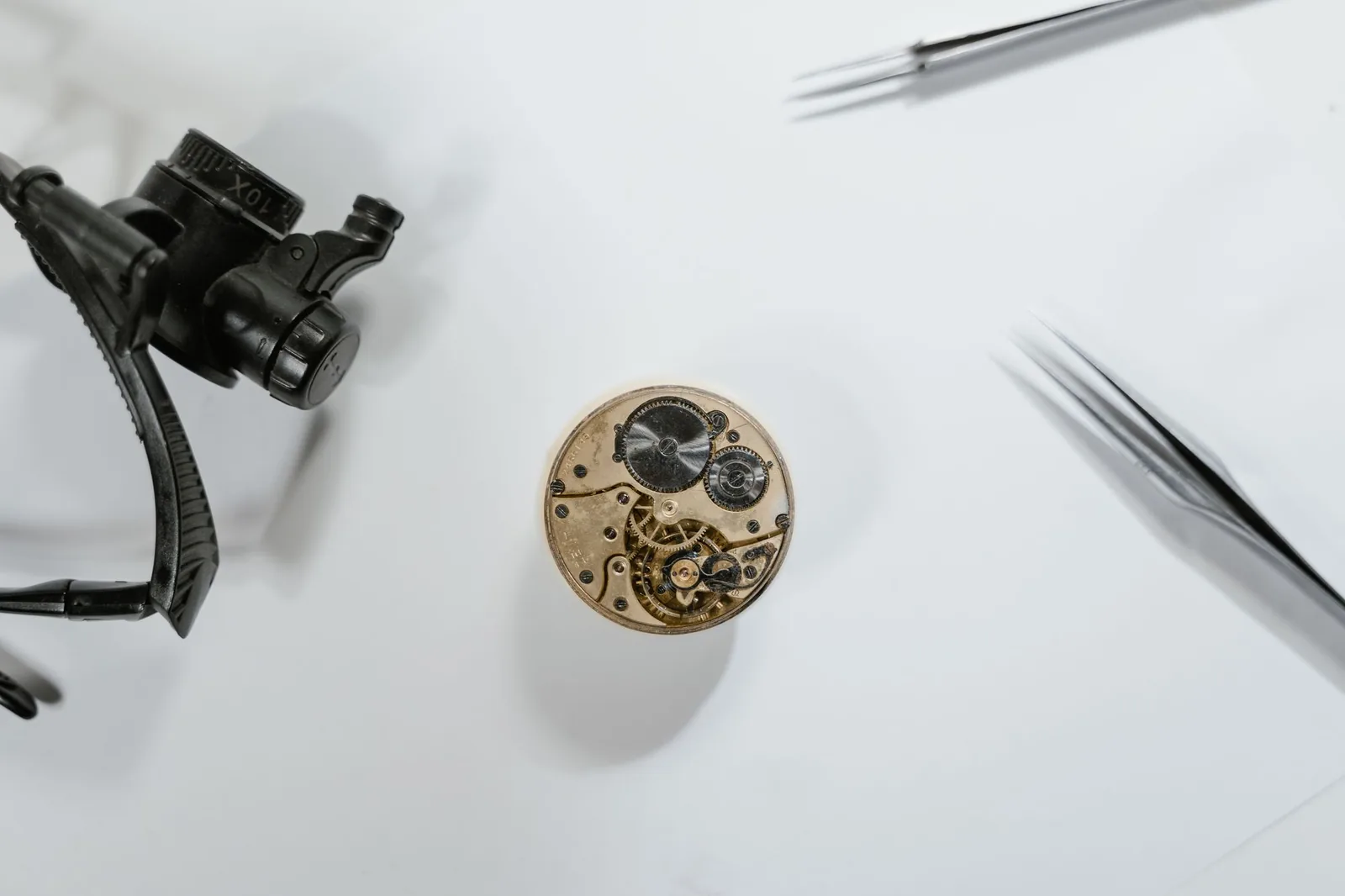 A bare watch movement on a bench beside a loupe and tweezers