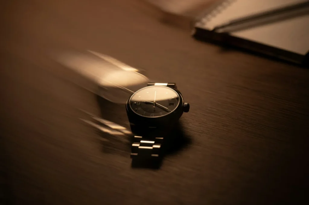 A steel wristwatch lying face up on a dark wooden table next to an open notebook and a pen